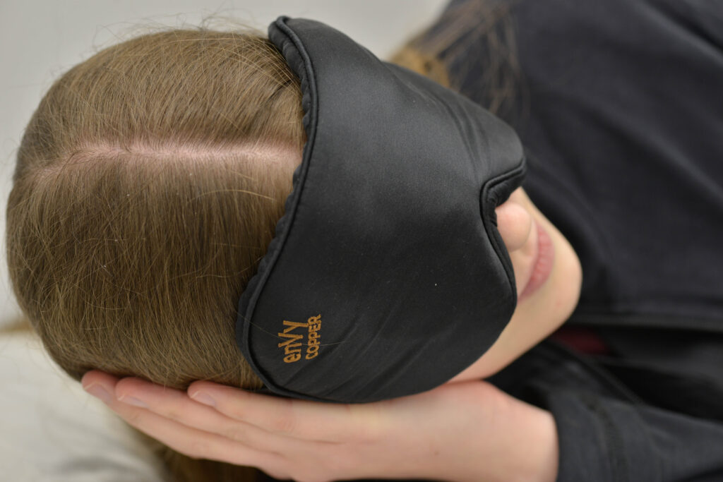 Close up image of a woman lying on her side with her hand under her head and wearing an enVy Copper silk eye mask.